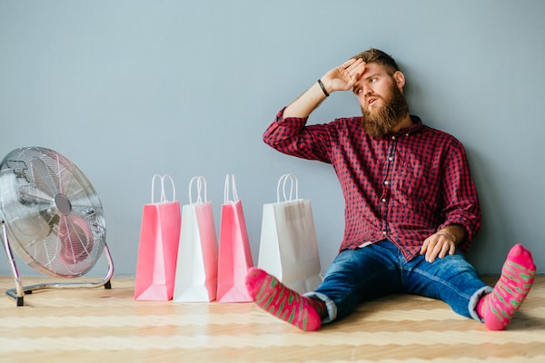 Portrait complet d'un hipster fatigué, barbu, en chaussettes colorées, jeans et chemise assis sur le sol avec des sacs à provisions roses et blancs sous fond de mur gris. Concept de la méthode CAB qui a bien fonctionné